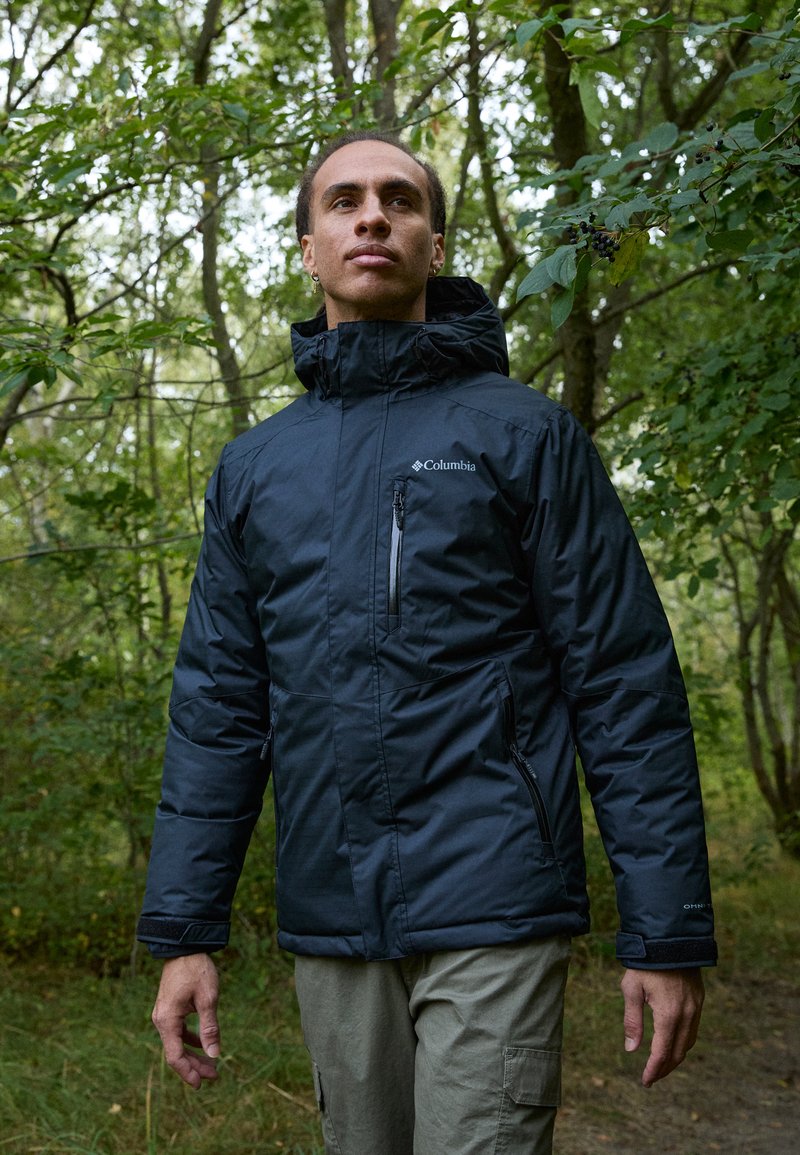 Schwarze isolierte Jacke mit einem mit Reißverschluss versehenen Brusttasche, hohem Kragen und verstellbarem Kapuze, getragen über hellfarbigen Cargo-Hosen in einem Waldsetting.