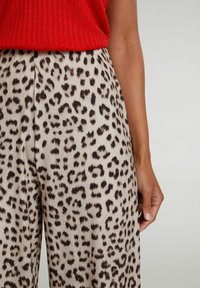 Kvinde i leopardmønstrede bukser iført en rød ribbet ærmeløs top, med hånden afslappet ved sin side foran en ensfarvet baggrund.