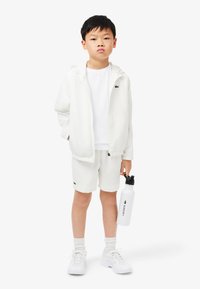 White lightweight jacket with hood, white t-shirt, and matching shorts; white sneakers and a white water bottle with black logo accents.
