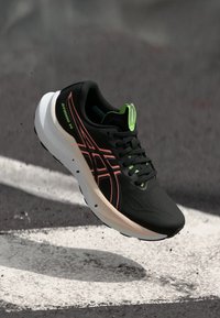 Ténis de corrida preto e verde néon com logótipo vermelho, em suspensão acima de alcatrão com marcações brancas, mostrando movimento e design leve.