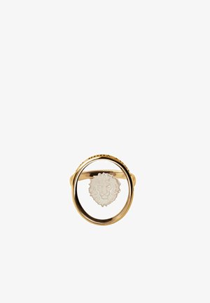 Anillo de oro con un marco circular y un emblema de cabeza de león blanco suspendido en el centro sobre un fondo blanco.