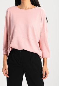 Un haut oversized rose clair avec un motif texturé, un col large et des manches trois-quarts, associé à un pantalon noir taille haute.