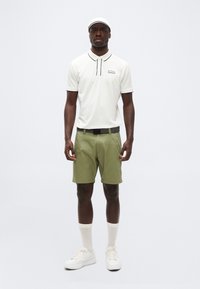 Άνδρας που φοράει λευκό καπέλο-σκιάστρο, λευκό πόλο με μαύρο περίγραμμα, χακί σορτς με μαύρη ζώνη, λευκές κάλτσες και λευκά αθλητικά παπούτσια, στέκεται αντιμετωπίζοντας προς τα εμπρός.