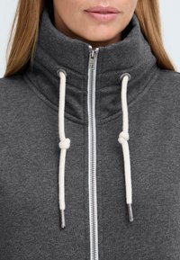 Grijze zip-hoodie met een hoge kraag, voorzien van witte trekkoorden, een zilveren rits en een zachte, gestucteerde stof.