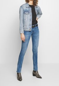 Person som bär en ljusblå denimjacka, svart skjorta, smala blå jeans med svärta, och klackstövlar med ormskinsmönster, står med en hand på höften.