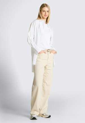 Blonde vrouw staat met handen in de zakken, draagt een wit hemd met knopen, een wit T-shirt, beige wijd uitlopende broek en witte sneakers tegen een effen achtergrond.