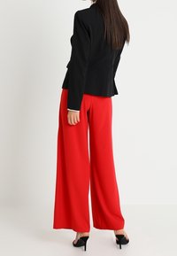 Kvinna klädd i en svart åtsittande blazer, vida röda byxor, svarta högklackade sandaler, stående med ryggen vänd framåt.