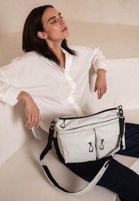 Borsa a tracolla grigia chiara con due tasche frontali con zip, cerniere nere e tracolla regolabile. Materiale morbido con una texture liscia.