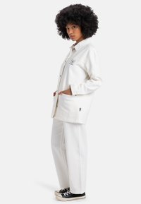 Veste surdimensionnée en coton blanc avec fermeture à boutons et deux poches avant, associée à un pantalon large assorti et des baskets noires.