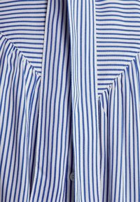 Chemise rayée avec des lignes verticales bleues et blanches, fermée par des boutons, avec des détails plissés et une texture de tissu léger.