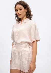 Γυναίκα που φοράει ανοιχτό ροζ μπλούζα με κοντά μανίκια και σχέδιο και ταιριαστό σορτς με ψηλή μέση, όρθια μπροστά σε απλό λευκό φόντο.