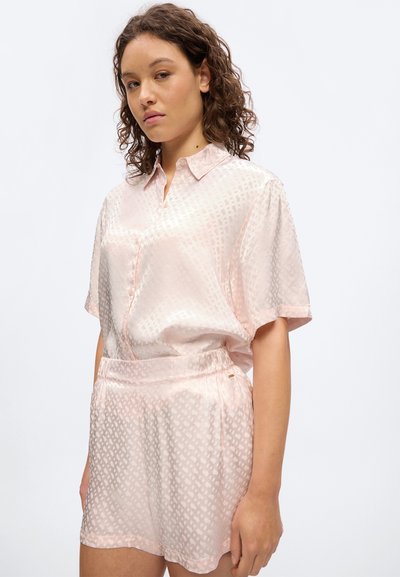 Femme portant un chemisier à manches courtes à motifs rose clair et un short taille haute assorti, debout devant un fond blanc uni.