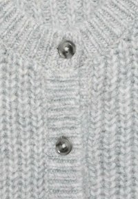 Sivi pleteni materijal s rebrastom teksturom. Ima dva velika, tamna dugmeta na izrezu, što dodaje kontrast i detalj.