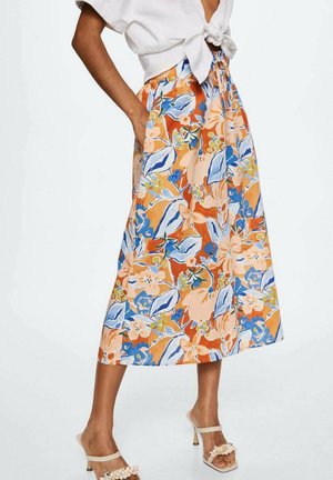 Frau trägt einen hoch taillierten Midirock mit Blumenmuster in Orange, Blau und Beige, kombiniert mit einer weißen gebundenen Bluse und beigefarbenen Sandaletten mit Absatz.