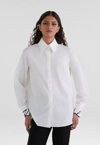 White button-up shirt with a classic collar, long sleeves, and black decorative stitching at the cuffs. Smooth fabric, relaxed fit.
