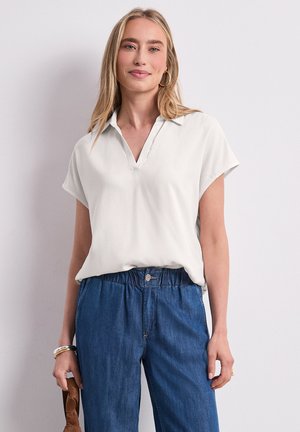 Femme portant un chemisier blanc à manches courtes avec col et un jean bleu élastique taille haute, debout devant un fond clair uni.