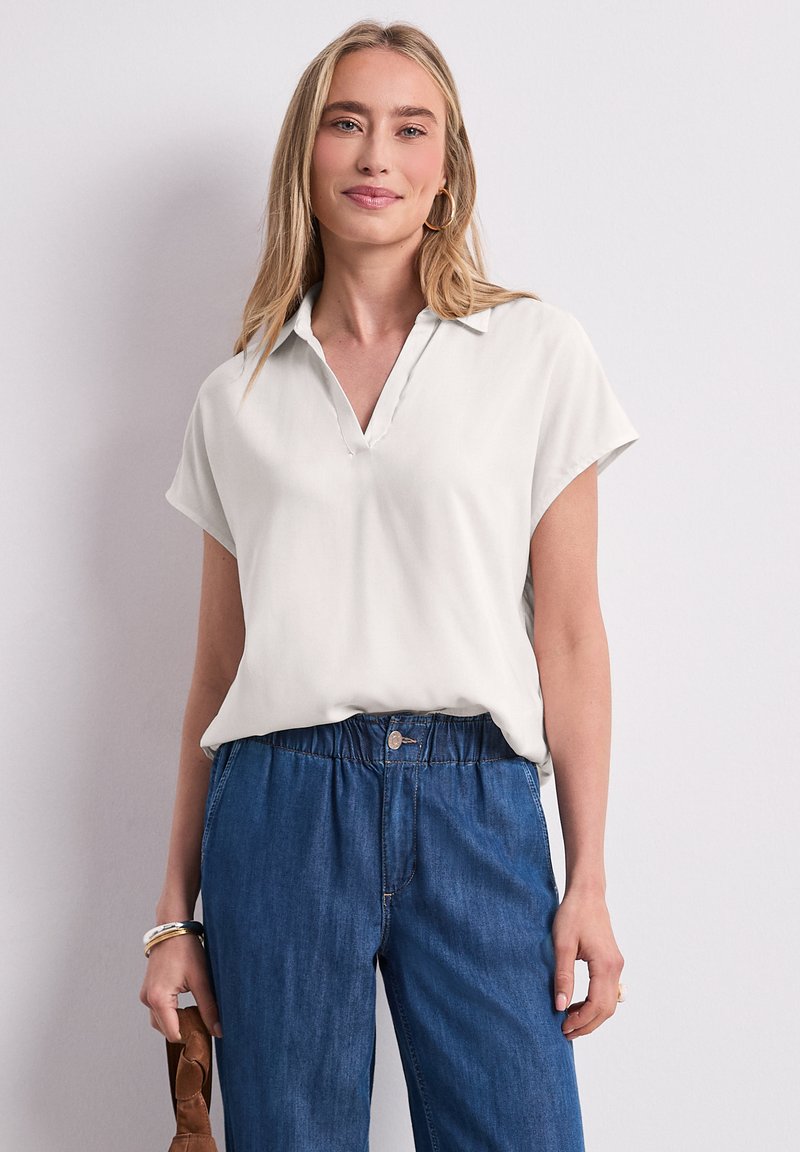 Femme portant un chemisier blanc à manches courtes avec col et un jean bleu élastique taille haute, debout devant un fond clair uni.