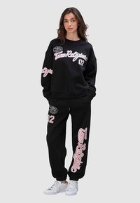Ensemble sweat-shirt noir et pantalon avec logo et numéros roses. Tissu semble doux, coupe décontractée, avec des détails textuels et graphiques visibles. Chaussures blanches.