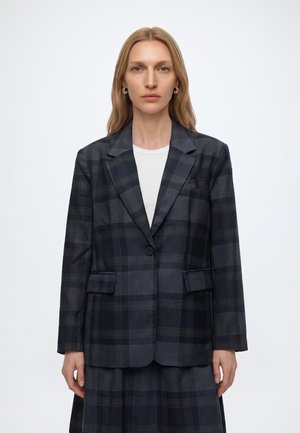 Femme aux longs cheveux blonds portant des boucles d'oreilles en or, un blazer à carreaux foncé et une chemise blanche, debout devant un fond uni.