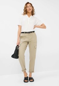 Beige cargobroek met opgerolde mouwen, een zwarte riem en een witte blouse met korte mouwen. Accessoires zijn een zwarte geweven handtas en zwarte sandalen.