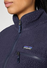 Navy fleece jacket with high collar, zippered front, and chest pocket; features textured fabric and Patagonia logo in multicolor detail.