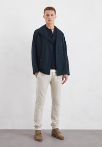 Mørkeblå dobbeltbrystet jakke, beige bukser, brune sko. Teksturen fremstår glat, med store revers og knapper. Modellen står foran en lys baggrund.