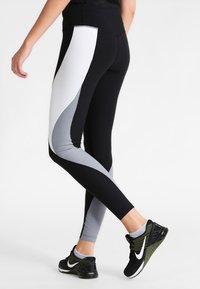 Person som bär svarta, vita och grå färgblockerade leggings och svarta Nike löparskor, som står med ett ben korsat bakom det andra.