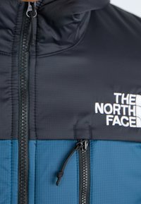 Blå och svart isolerad jacka med dragkedja framtill. Har strukturerad tyg, logotyp på bröstet och justerbar dragsko vid huvan.