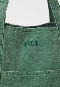 Zelena platnena torba s ušivenim džepom i izvezenim "GAP" logotipom na gornjem delu. Izrađena od izdržljivog materijala.