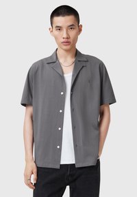 Camisa gris de manga corta, hecha de tela suave, que presenta un cuello, botones en el frente y un sutil detalle de logo en el lado izquierdo.