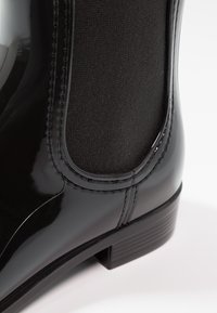 Bottine en cuir noir verni avec panneaux latéraux élastiques, bout arrondi et petit talon. Fini lisse avec tissu mat contrastant.