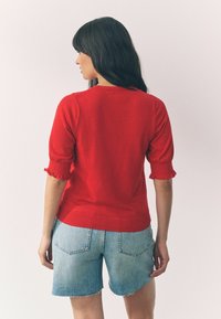 Pull rouge à manches courtes avec un volant froncé aux poignets, associé à un short en denim bleu clair avec un ourlet effiloché.