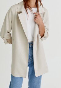 Jasnobeżowa marynarka o luźnym kroju, z klapami, podwiniętymi rękawami i bocznymi kieszeniami, noszona na białym t-shircie i niebieskich dżinsach.