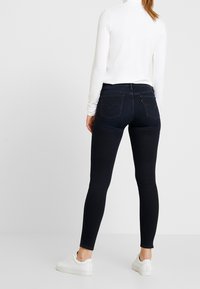 Jeans oscuros ajustados de tiro alto, con bolsillos traseros que tienen un patrón de costura, combinados con una camiseta blanca de manga larga y zapatos blancos.