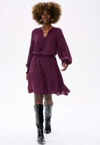 Pruimen jurk met een v-hals, lange mouwen en een gegatherde rok. Gecombineerd met zwarte leren kniehoge laarzen, een gladde textuur en een subtiel patroon.