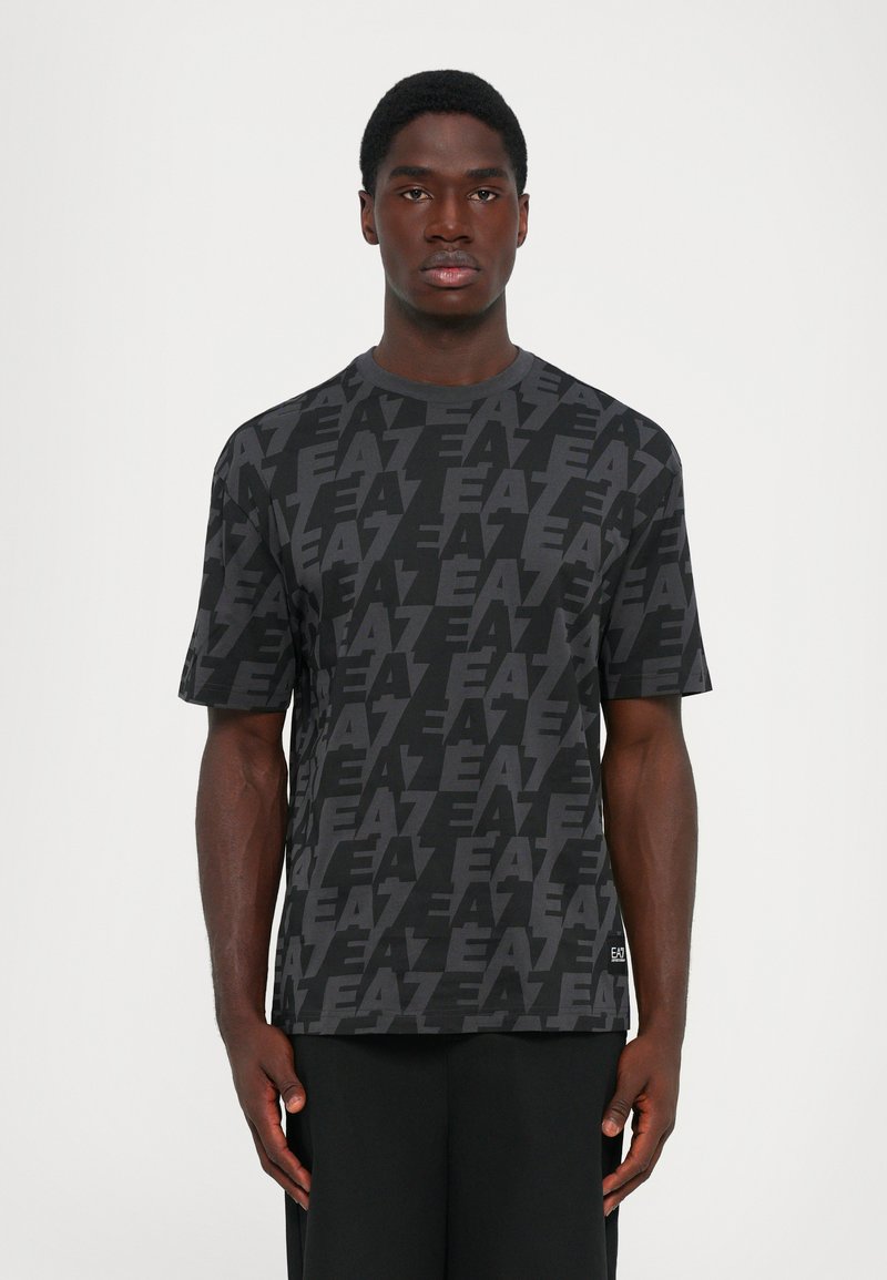 Man wearing black and dark gray patterned EA7 logo t-shirt, standing with arms relaxed against a plain white background.