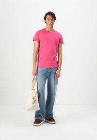 Homme portant un t-shirt rose, un jean bleu ample et des baskets marron, tenant un sac fourre-tout beige, se tenant devant un fond blanc.