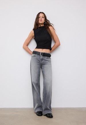 Femme portant un haut noir sans manches, un jean gris à jambes larges avec ceinture noire, et des chaussures noires, debout contre un mur blanc uni.