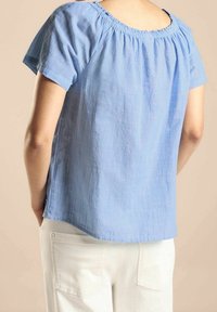 Jasnoniebieska bluzka z krótkim rękawem z marszczoną linią dekoltu, wykonana z teksturowanej tkaniny, charakteryzująca się luźnym krojem i zaokrąglonym hem.