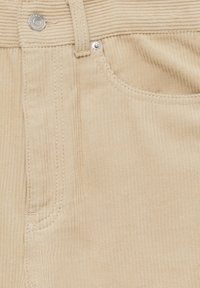 Beige corduroy stof met verticale ribbels, voorzien van een voorzak, metalen knopen en stiksels langs de naden.
