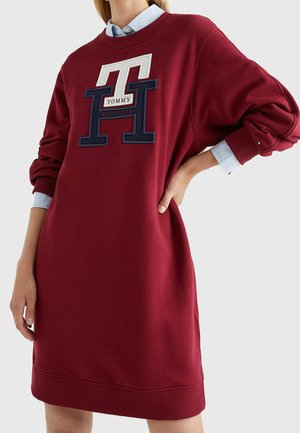 Kobieta ubrana w długą czerwoną sukienkę typu sweatshirt z prążkowanymi mankietami i dużym logo Tommy Hilfiger na piersi, nałożoną na niebieską koszulę z kołnierzykiem.