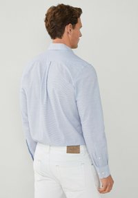 Camicia a maniche lunghe a righe blu chiaro, in cotone, con polsini a bottone, yoke posteriore, abbinata a pantaloni bianchi con etichetta del marchio color sabbia.
