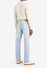 Jeans de pierna recta en azul claro con un parche de cuero en la cinturilla, complementados por una sudadera beige y zapatillas blancas con suelas de goma.