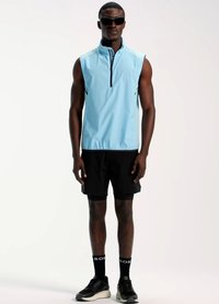 Hellblaue ärmellose Jacke mit hohem Kragen und Reißverschlussdetails, kombiniert mit schwarzen Shorts und schwarzen Sneakers. Schwarze Socken mit weißem Logo.