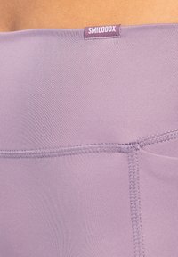 Leggings viola con una texture liscia e elastica. La fascia in vita presenta un piccolo etichetta con la scritta "SMILOOOX" e dettagli di cucitura visibili.