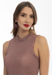 Rózsaarany, ujjatlan felső, magas nyakkivágással, csillogó textúrával. Kerek rózsaszín fülbevalók ellentétes részletként jelennek meg.