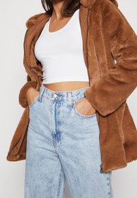 Brauner Kunstpelzmantel mit Kapuze, Reißverschluss und Seitentaschen, getragen über einem weißen Crop-Top und hellblauen Jeans mit verblasster Textur.