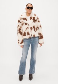 Casaco cropped de pele sintética com padrão de vaca castanho e branco, com bolsos frontais, usado sobre uma camisa branca, combinado com jeans flare azuis e botas escuras.