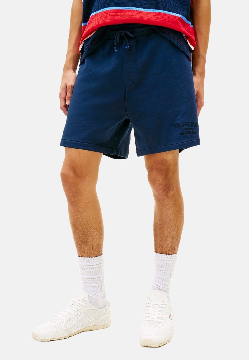 Homme portant un short bleu marine à cordon, des chaussettes blanches, des baskets blanches et un haut bleu avec des rayures rouges et blanches.