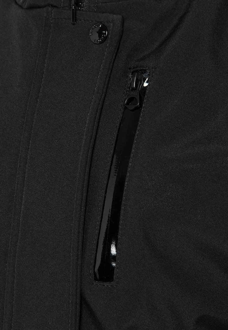 Veste noire en tissu texturé, avec une poche zippée dotée de quincaillerie noire brillante et une finition lisse au niveau du bord de la fermeture éclair.